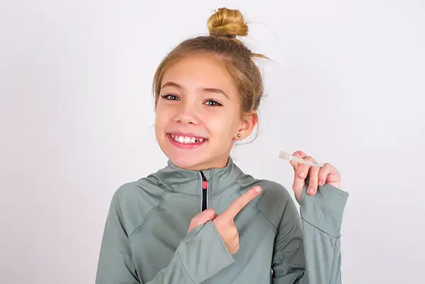 Young girl in a gray sweatshirt smiling and pointing to a clear aligner she is holding.