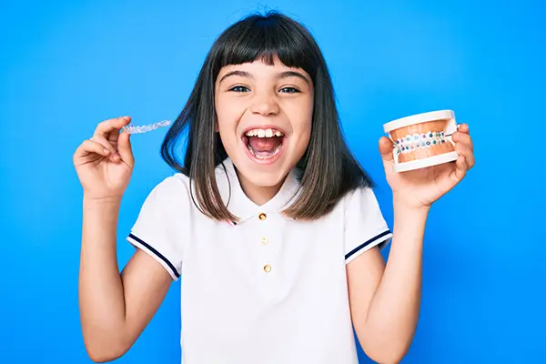 Ready to begin her orthodontic treatment, a young girl excitedly holds up clear aligners in one hand and a dental model with traditional braces in the other hand.