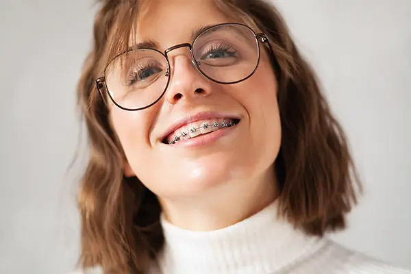 A young woman with glasses smiling brightly, highlighting her traditional metal braces.